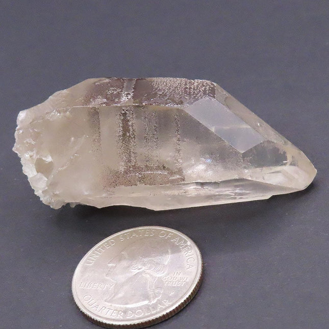 Silver Smoky Lemurian Seed Quartz Crystal Point with Time-Link & Rainbow next to a U.S. quarter dollar on a gray background